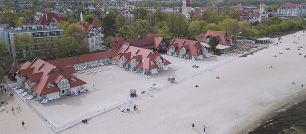 Vista aérea de una playa con varios edificios de techos rojos, algunos con balcones, y una zona de playa arenosa con algunas personas.