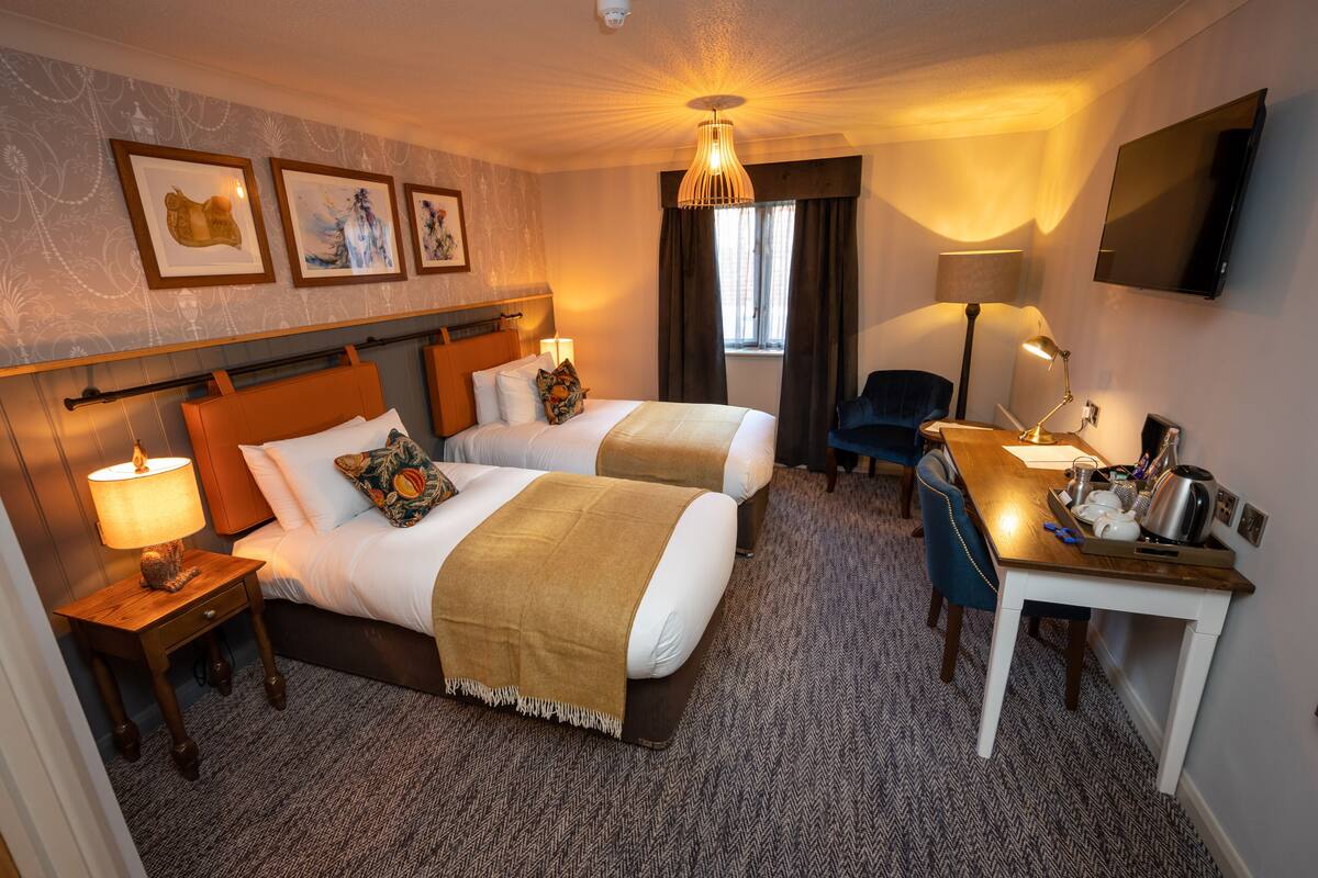 A hotel room with two beds, a desk, a chair, a TV, and artwork on the wall.