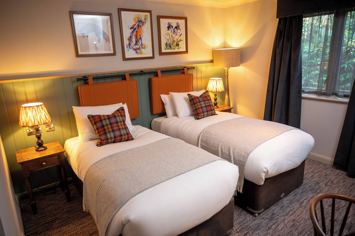 A hotel room with two beds, a nightstand with a lamp, a window with curtains, and artwork on the wall.