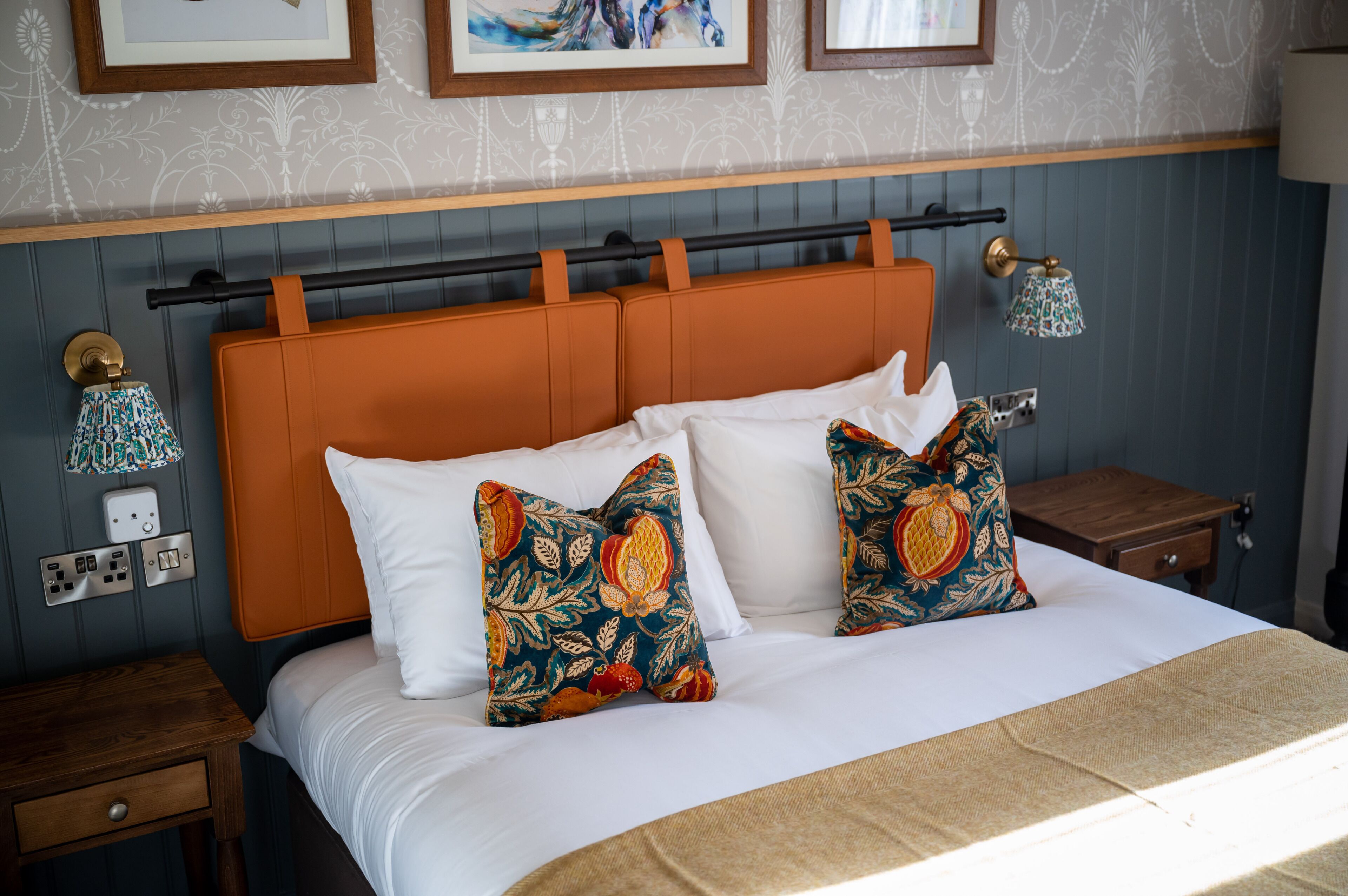 A neatly made bed with white linens and two decorative pillows, a wooden nightstand with a drawer, and a wall-mounted lamp.