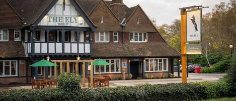 A traditional Tudor-style pub named "The Ely" with a signboard, outdoor seating, and a well-maintained hedge in front.