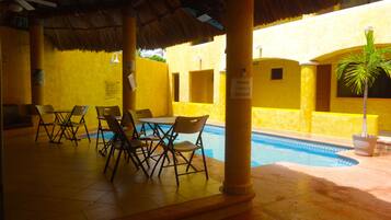 Uma área interna com piscina, paredes amarelas, teto de palha e um espaço para sentar com cadeiras e mesas.