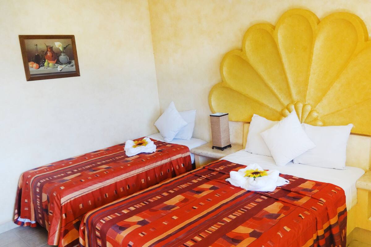 Una habitación de hotel con dos camas, un cabecero grande y decorativo, una mesita de noche pequeña con una lámpara, y un cuadro en la pared.
