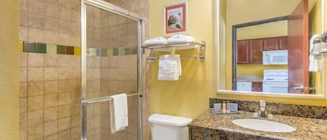 A bathroom with a glass shower enclosure, a toilet, a sink with a granite countertop, and a mirror with lights above.