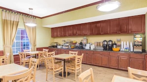 Un comedor con mesas y sillas de madera, una encimera de cocina con distintos electrodomésticos y una ventana con cortinas.