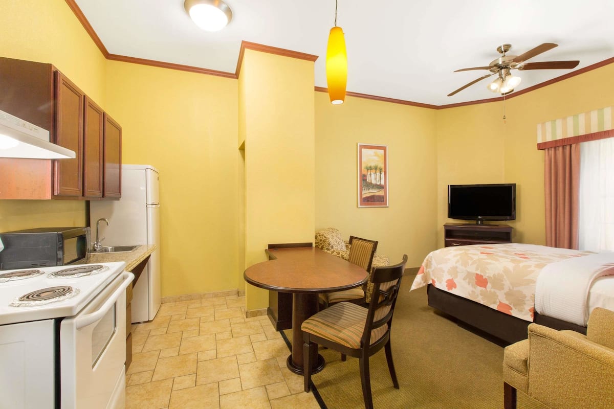A compact hotel room with a kitchenette, a dining table with chairs, a bed, a TV, and a ceiling fan.
