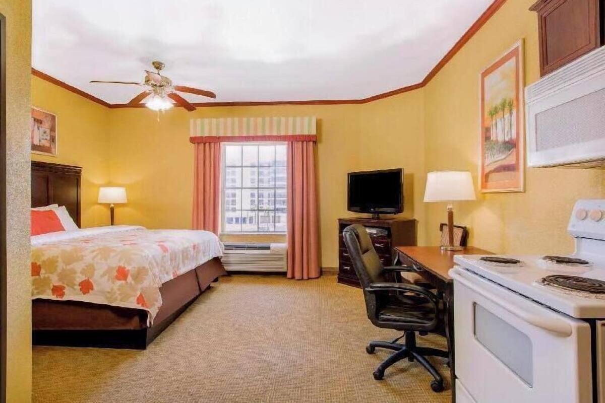 A hotel room with a bed, a desk with a television, a chair, an oven, and a window with curtains.