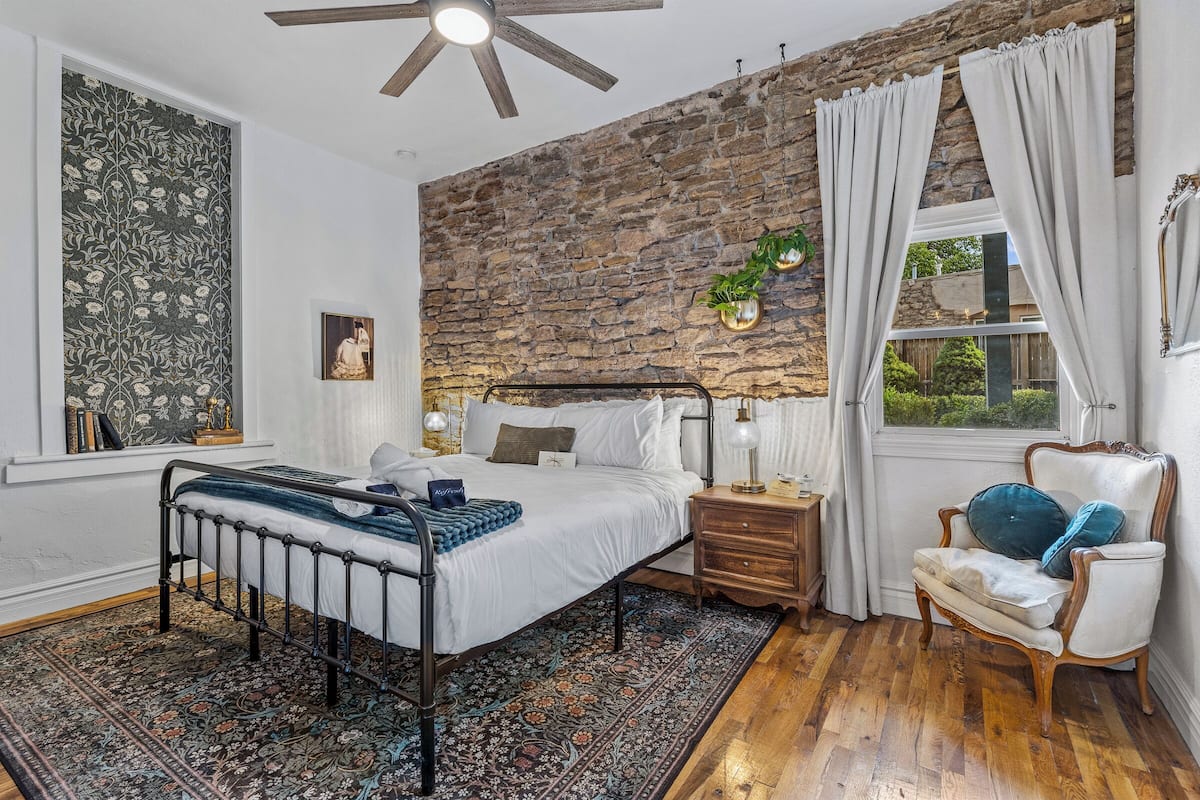 Une chambre avec un mur en pierre, un lit avec une tête de lit, une table de chevet, une chaise et une fenêtre avec des rideaux.