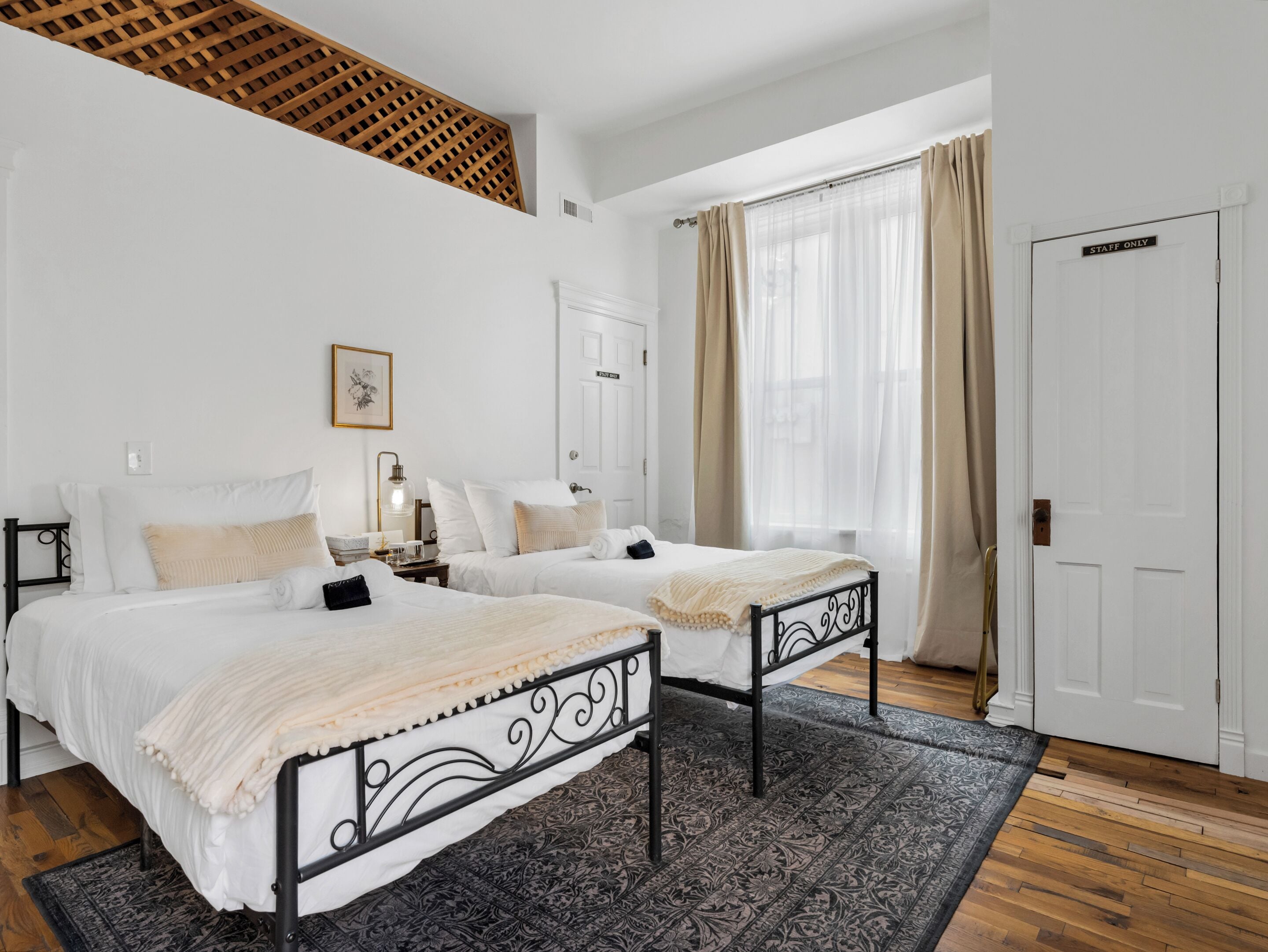 A hotel room with two beds, a wooden ceiling, a large window with curtains, and a 'STAFF ONLY' door.