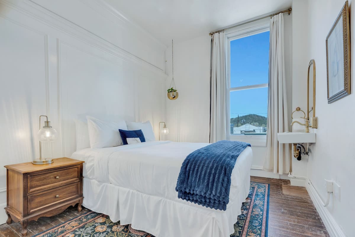 Une chambre avec un grand lit, une table de chevet, une lampe de chevet, une fenêtre avec vue et un lavabo fixé au mur.