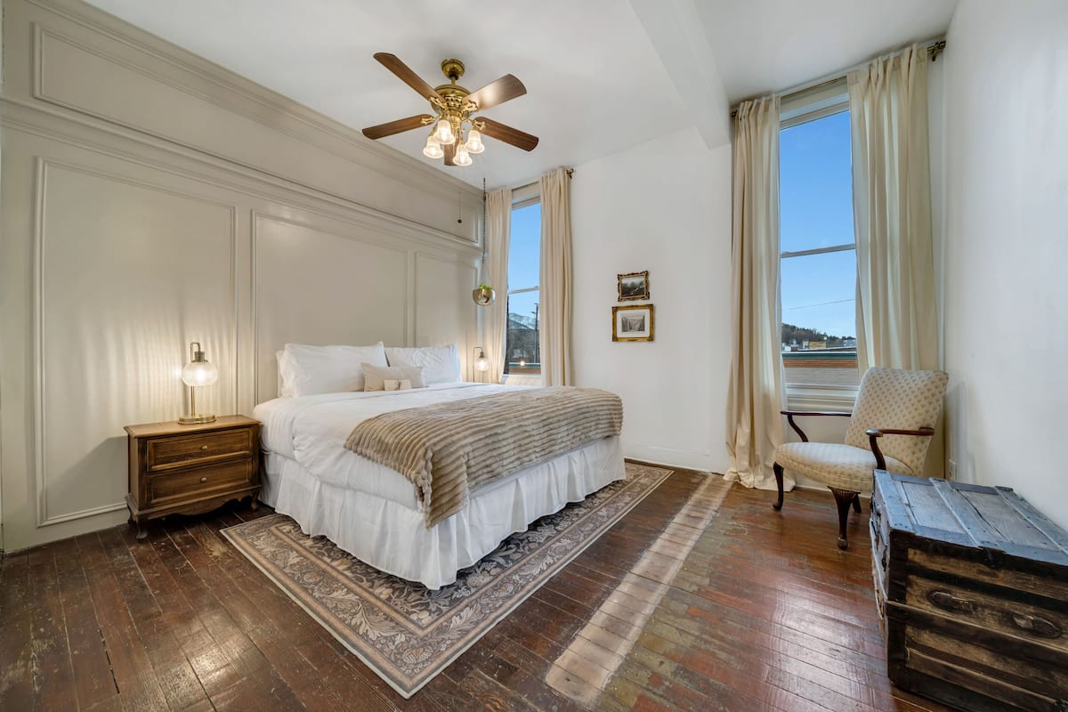 Une chambre avec un grand lit, des tables de chevet en bois, un ventilateur de plafond, une fenêtre avec des rideaux et un fauteuil.