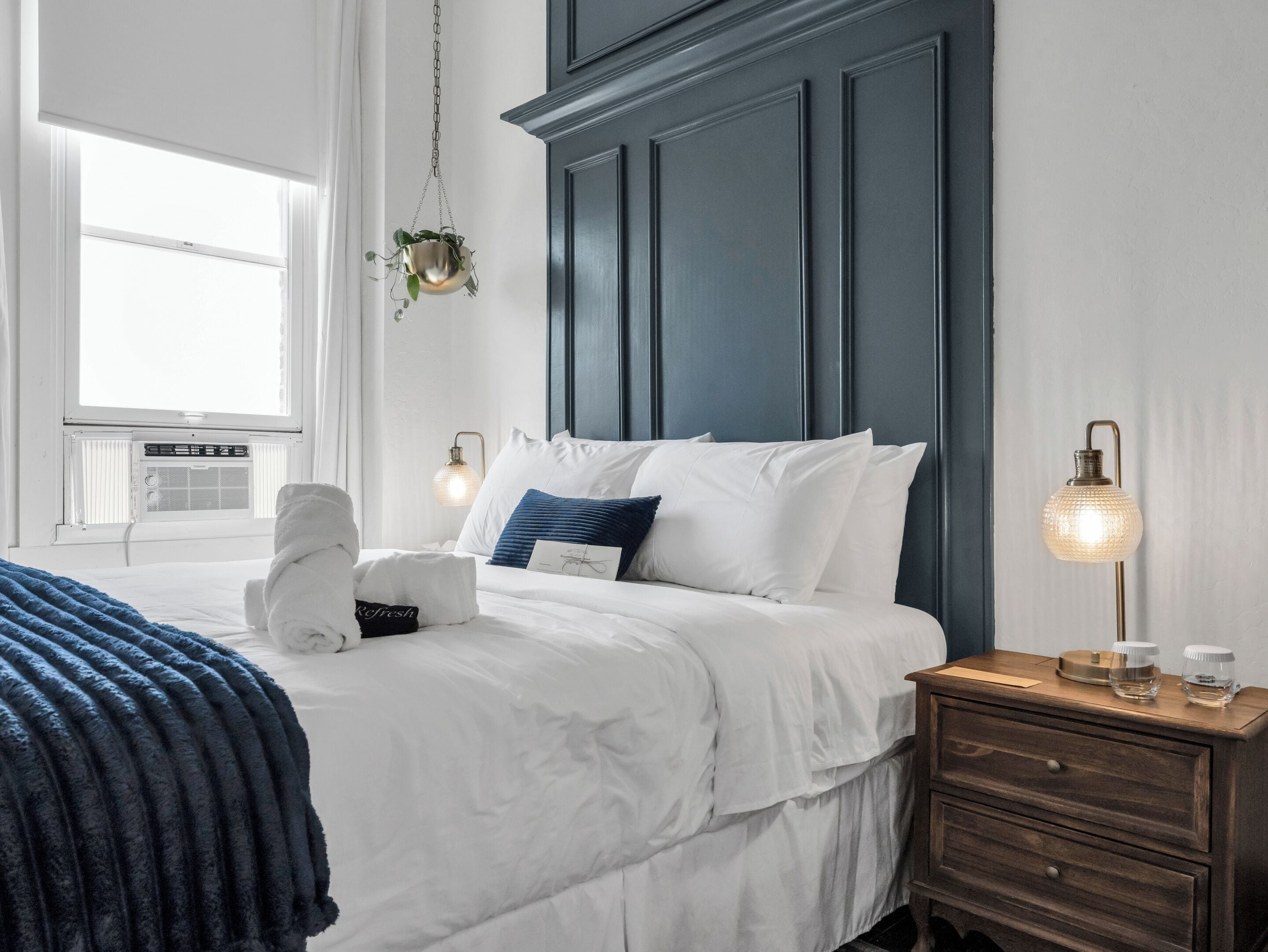 Un dormitorio con cama, mesitas de noche, una planta colgante y una ventana con persianas.