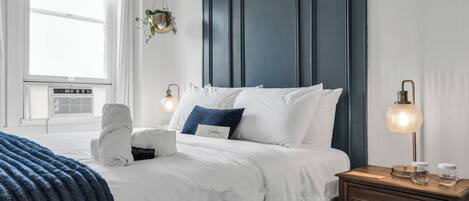 Un dormitorio con cama, mesitas de noche, una planta colgante y una ventana con persianas.
