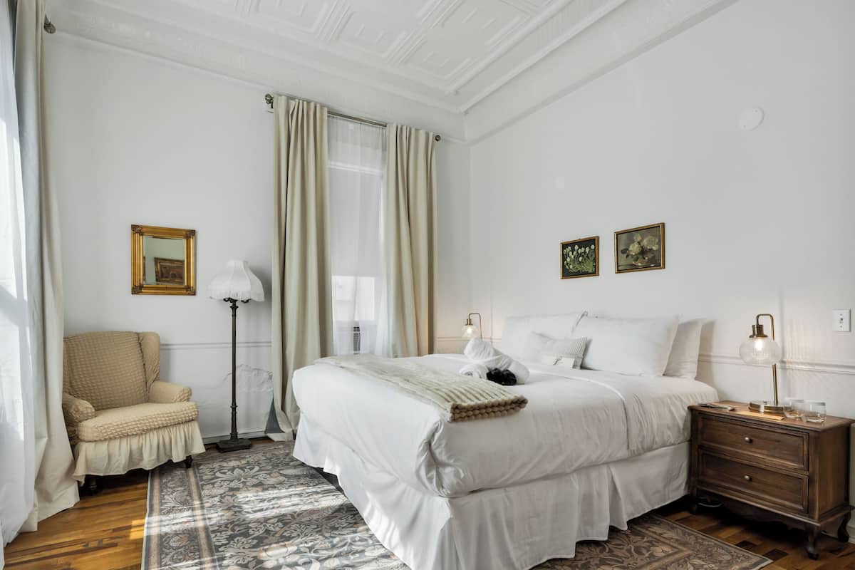 Une chambre à coucher avec un lit, une chaise, une table de chevet, une lampe et une fenêtre avec des rideaux.