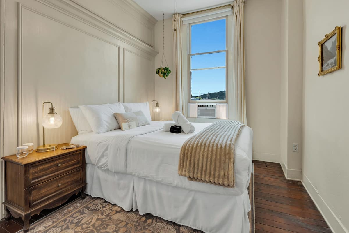 Une chambre à coucher avec un grand lit, une table de chevet en bois, une lampe sur pied et une fenêtre avec des rideaux.