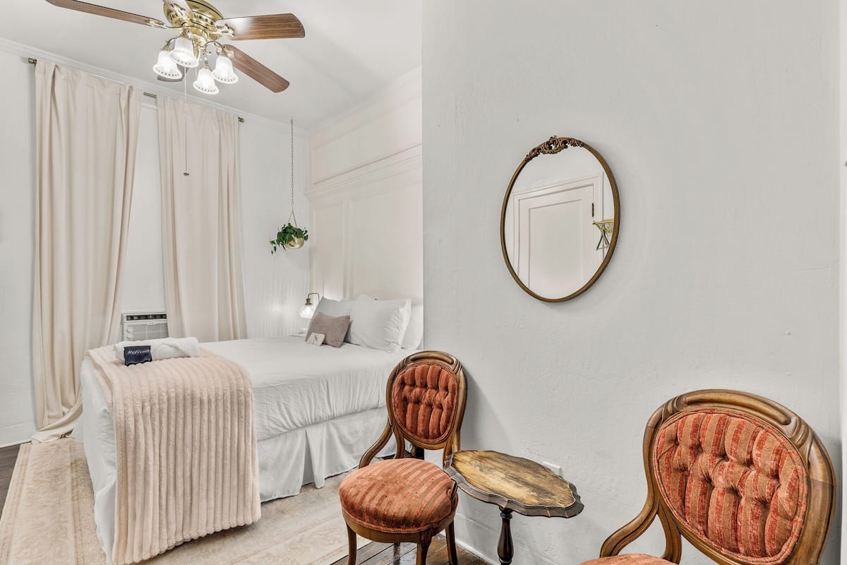 Une chambre à coucher avec un lit, une chaise, une table, un ventilateur de plafond et un miroir au mur.