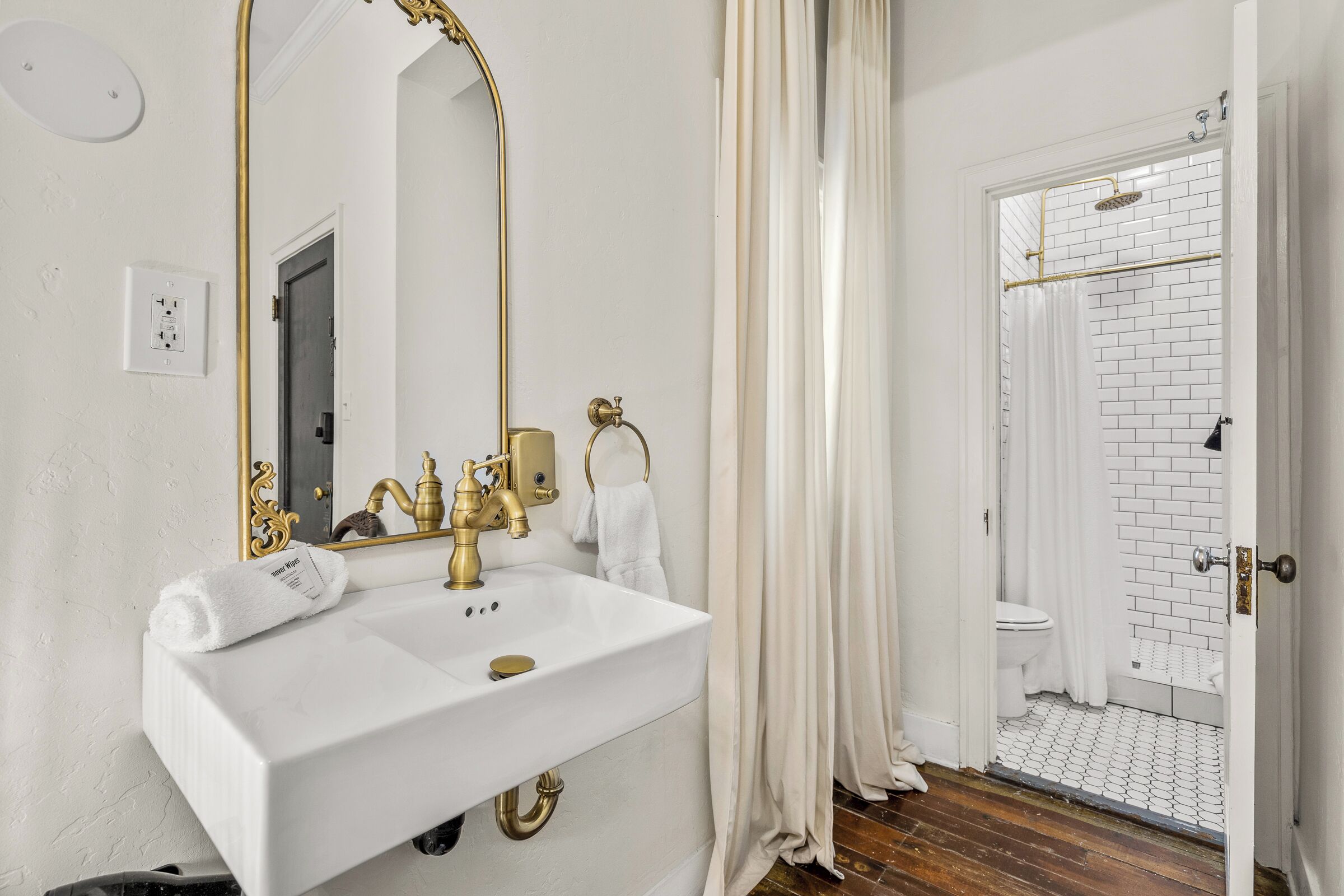 Un baño con lavamanos blanco, detalles dorados, un espejo grande y una zona de ducha con azulejos blancos y suelo de madera.