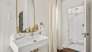 Un baño con lavamanos blanco, detalles dorados, un espejo grande y una zona de ducha con azulejos blancos y suelo de madera.