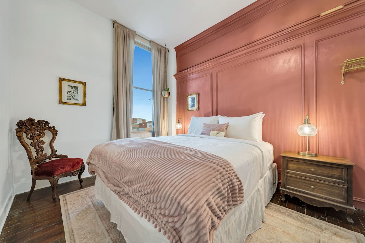 Une chambre à coucher avec un grand lit, une table de chevet en bois, un fauteuil rembourré de couleur rouge et une fenêtre avec des rideaux.