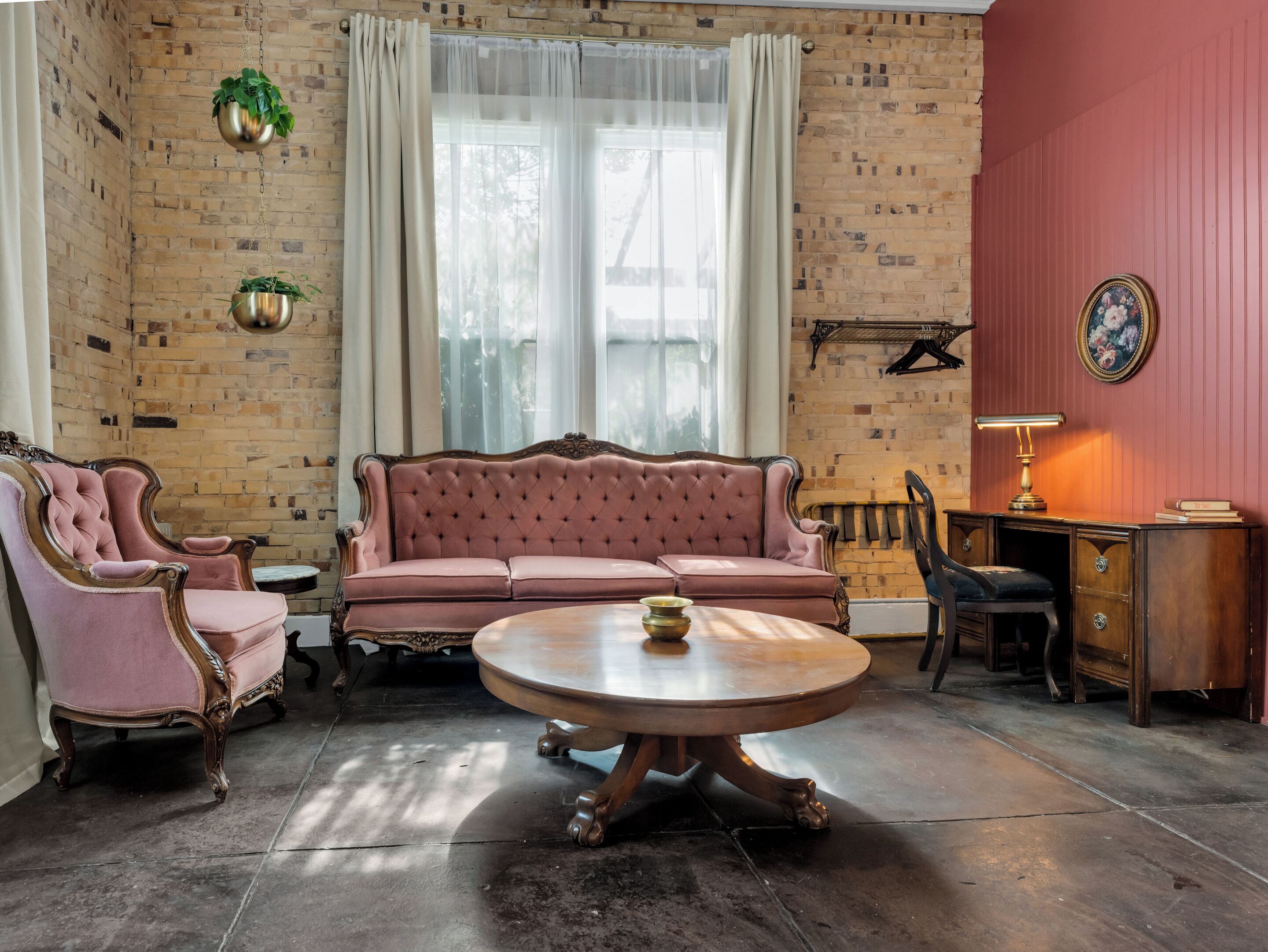 A room with a brick wall, a pink sofa, a round wooden table, and a desk with a lamp.