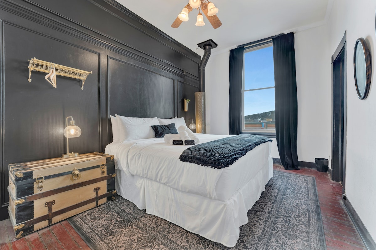Un lit bien fait, avec du linge de lit blanc, une tête de lit sombre et un coffre décoratif placé à côté.