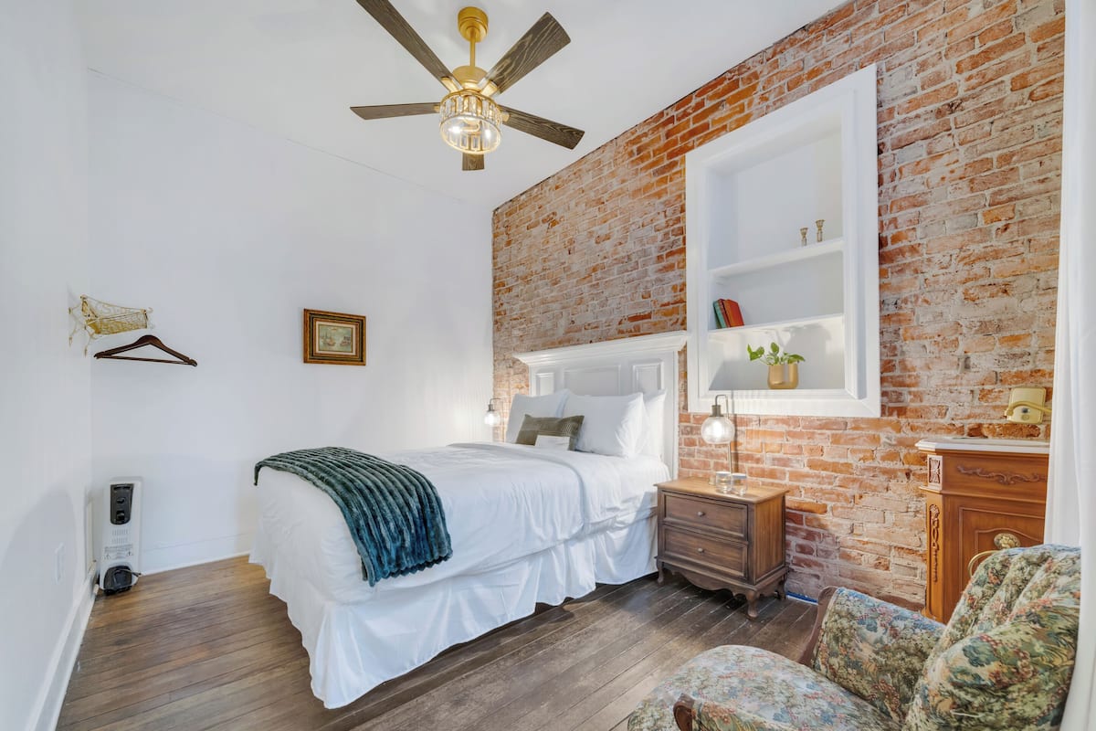 Une chambre avec un mur en briques, un lit avec une literie blanche, un plancher en bois, un ventilateur de plafond, une table de chevet avec une lampe et une chaise.