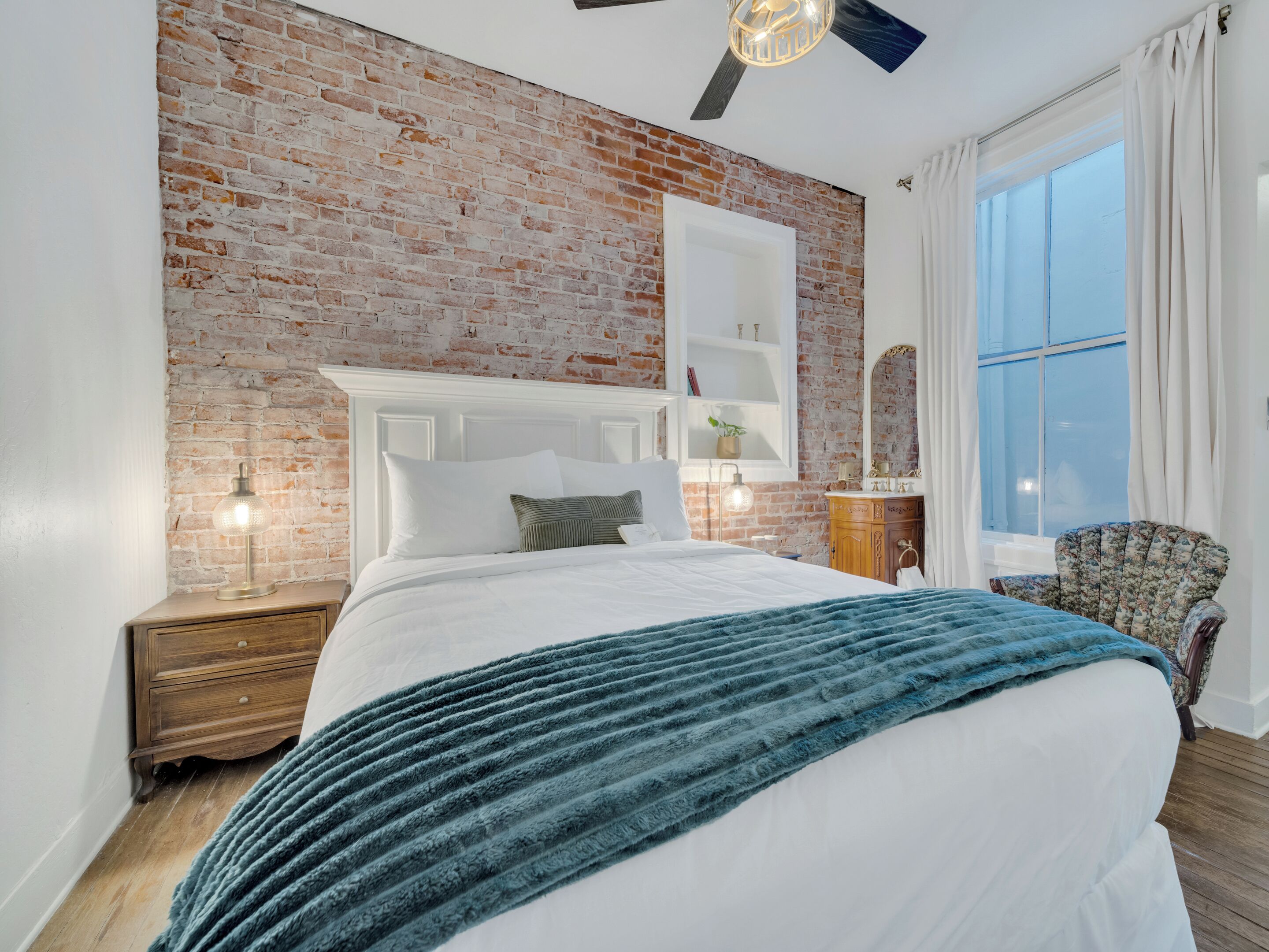 Une chambre avec un mur en briques, une tête de lit blanche, un grand lit recouvert d’une couverture bleue, une table de chevet en bois, une petite table avec une lampe, une chaise et une fenêtre avec des rideaux.