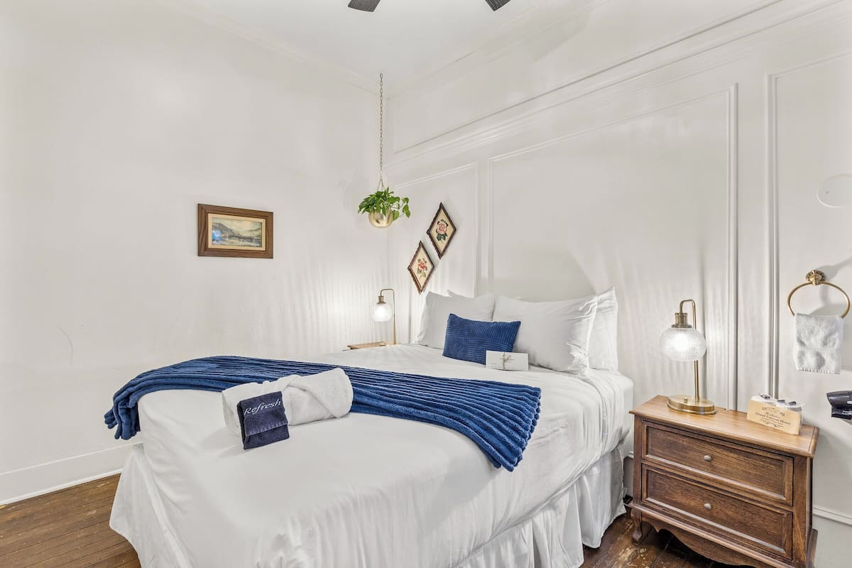 Un lit bien fait, avec du linge de lit blanc et des touches de bleu, une table de chevet avec une lampe et une plante suspendue.