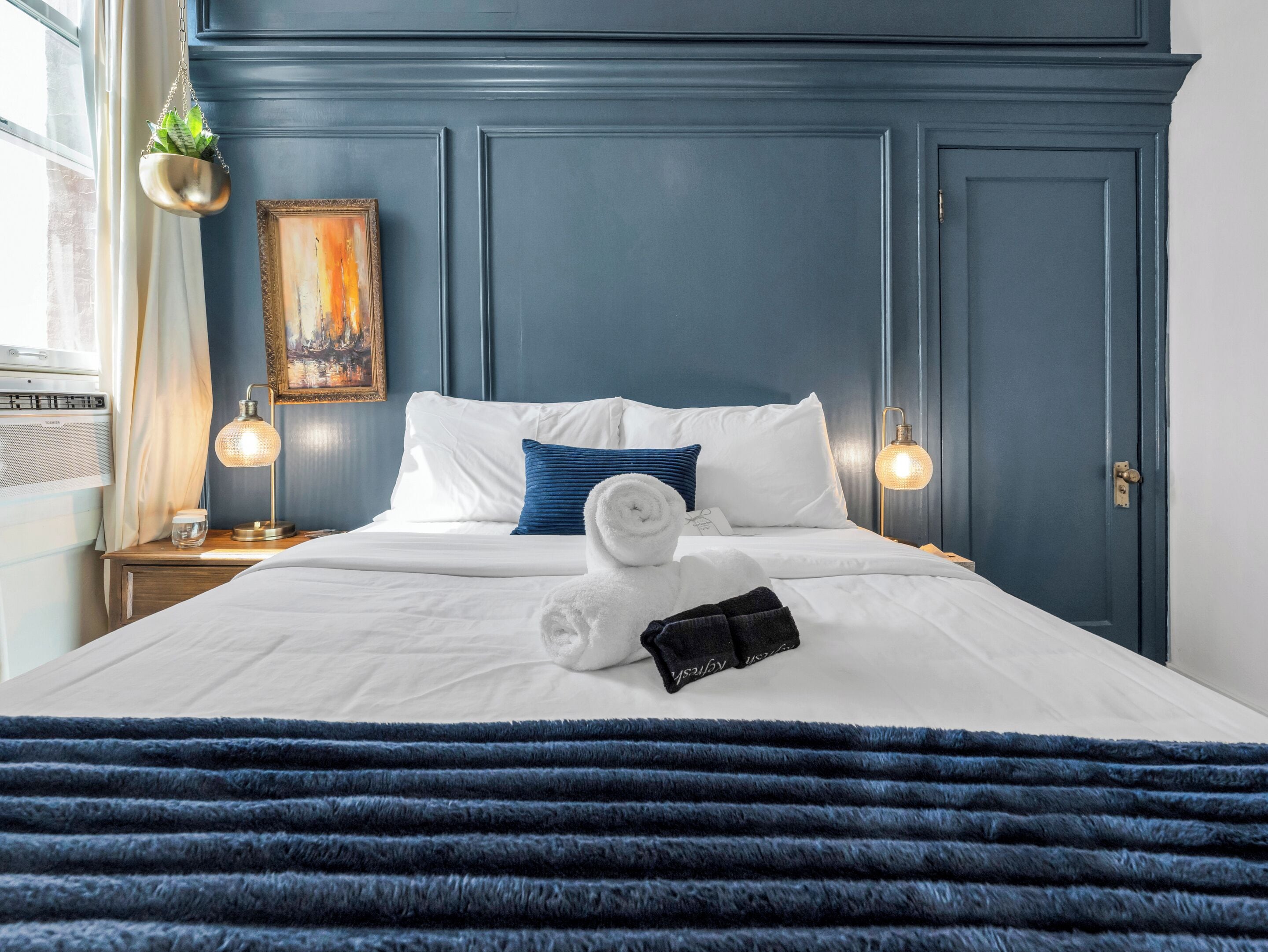A neatly made bed with white linens, a blue striped pillow, and a folded towel. There is a small potted plant hanging from the ceiling, a framed artwork on the wall, and a bedside lamp on the nightstand.