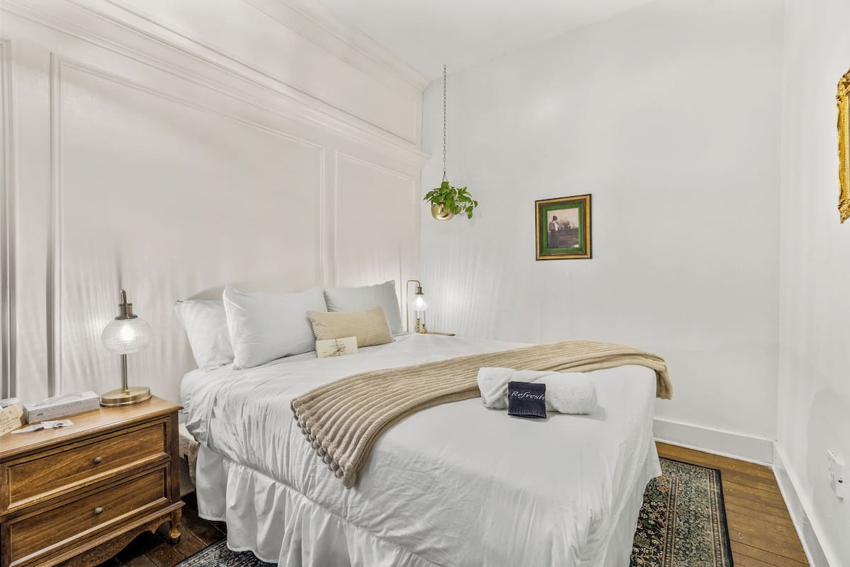 Une chambre à coucher bien rangée, avec un grand lit, des tables de chevet, une plante suspendue et un tableau encadré au mur.