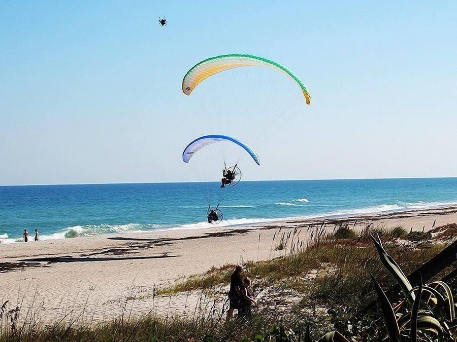 Een strand met drie paragliders in de lucht, een zandstrand en een helderblauwe lucht.