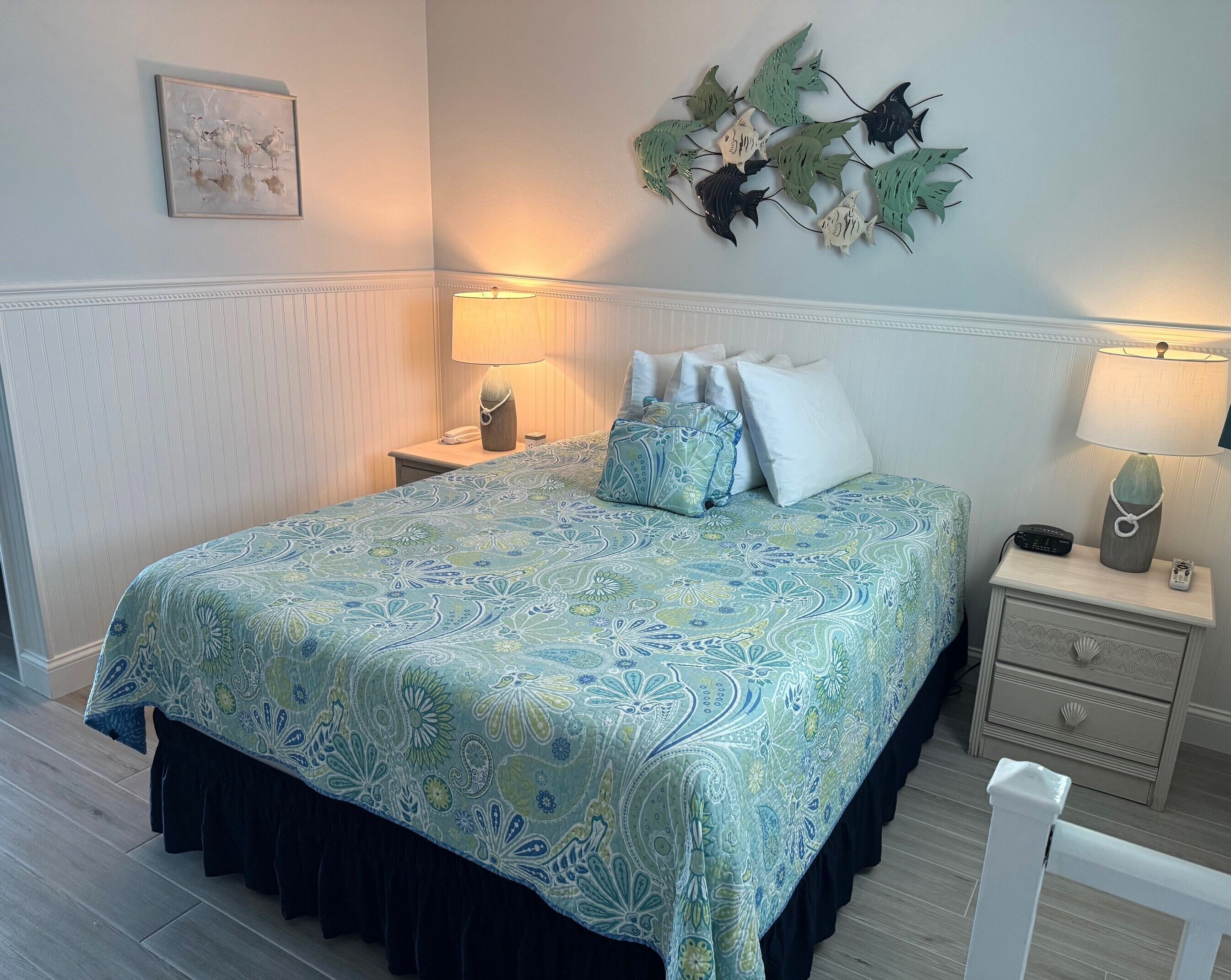 A neatly made bed with a patterned bedspread, two bedside lamps, a wall-mounted artwork, and a decorative leaf arrangement.
