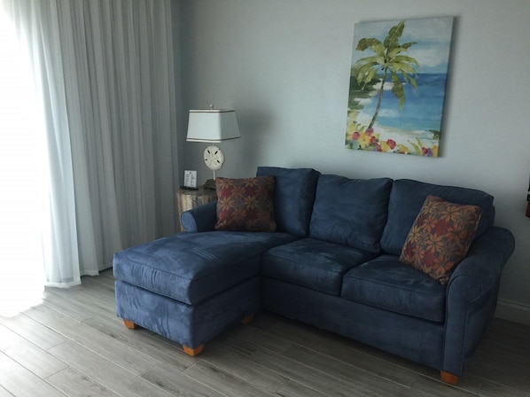 A living room with a blue sectional sofa, a side table with a lamp, a clock, and a painting of a beach scene.
