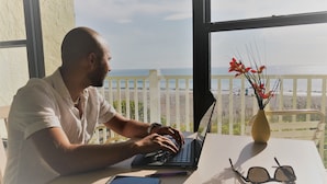 Una persona trabajando en un ordenador portátil a una mesa con vista a la playa y al océano.