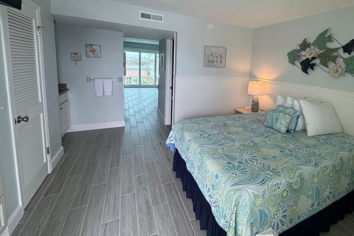 A bedroom with a bed, a nightstand, a lamp, a door, and a hallway leading to an outdoor area.