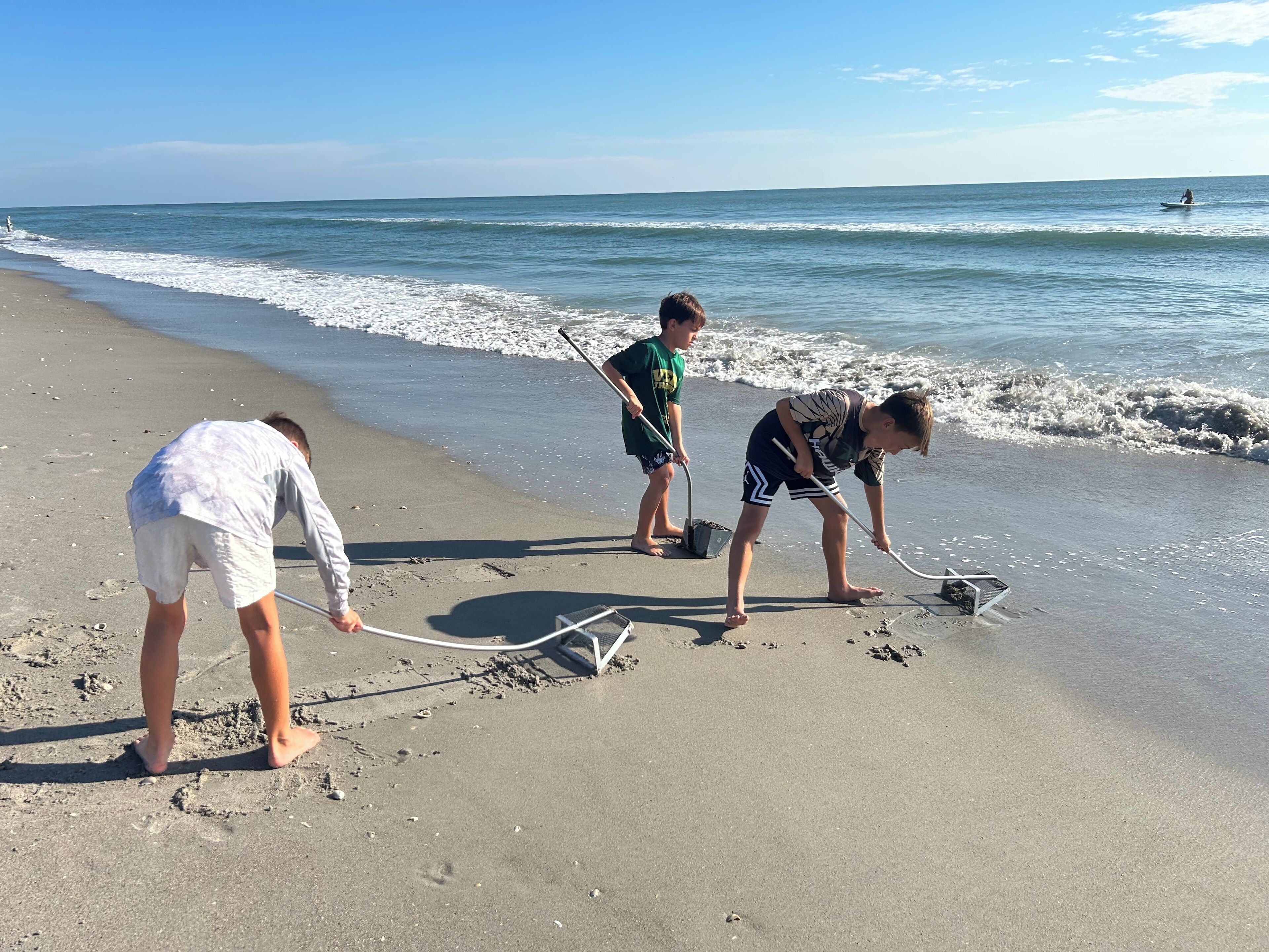 Drei Personen am Strand, eine mit einem Rechen und eine andere mit einem Netz, mit dem Meer im Hintergrund.