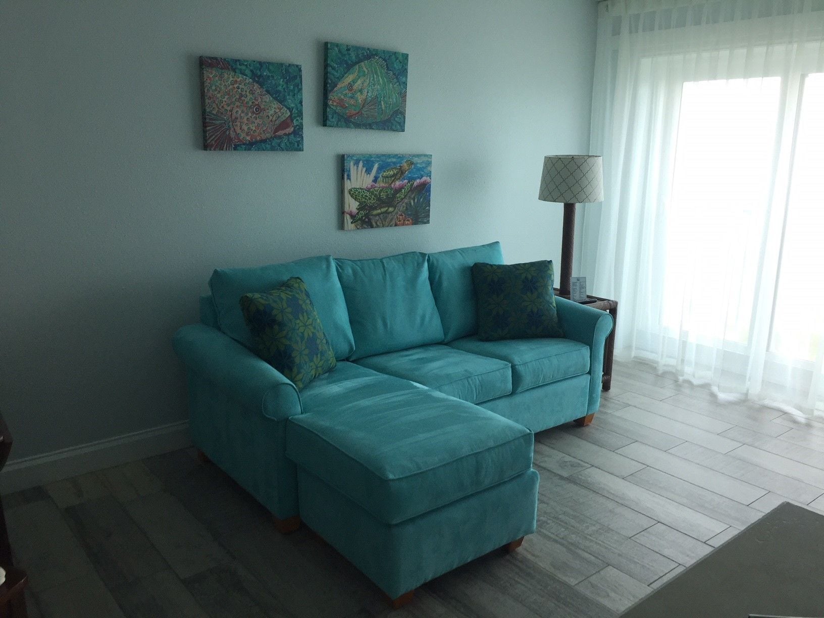 A teal-colored sofa with matching cushions in a room with light-colored wooden flooring. There are three wall-mounted paintings depicting marine life. A floor lamp with a patterned lampshade is positioned near a window with sheer white curtains.
