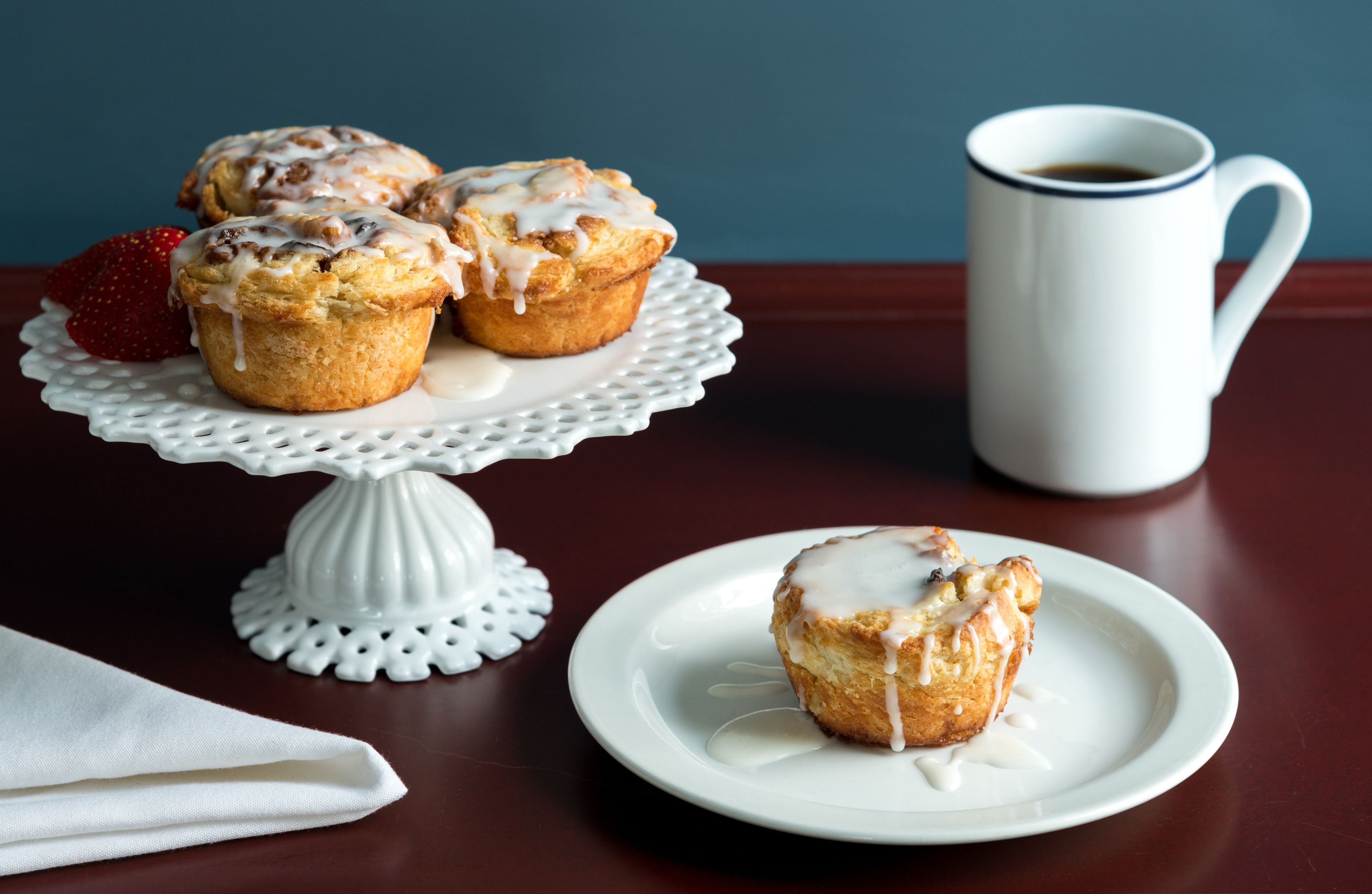 Ein Tisch mit einem weißen Kuchenständer, der drei Muffins hält, ein Teller mit einem Muffin, eine weiße Tasse und ein gefaltetes weißes Servietttuch.