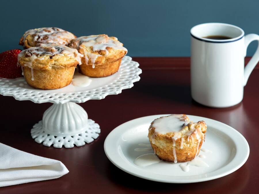 Ein Tisch mit einem weißen Kuchenständer, der drei Muffins hält, ein Teller mit einem Muffin, eine weiße Tasse und ein gefaltetes weißes Servietttuch.