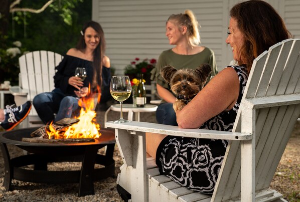A group of people sitting around a fire pit with a dog, in an outdoor setting with chairs and a table.
