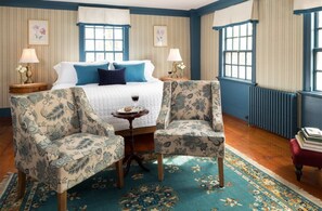 A room with a bed, two armchairs, a small table with a wine glass, and a red ottoman.