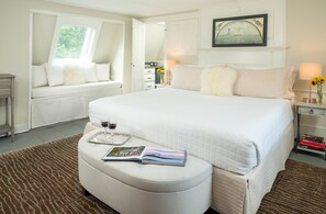 A bedroom with a large bed, a window seat, a bedside table with a lamp, and a small table with a tray and a book.