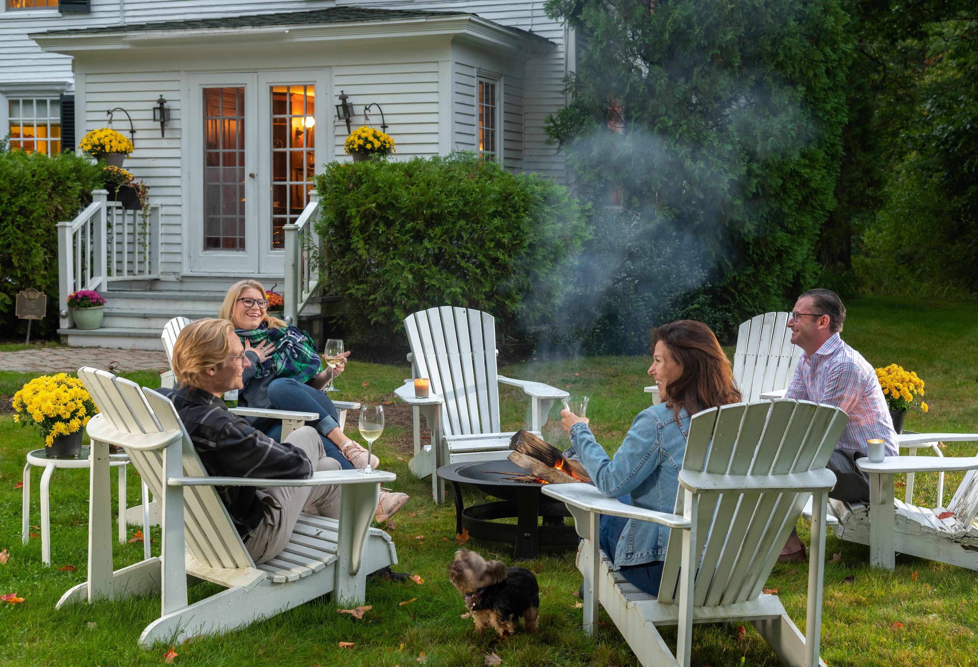 A group of people seated on white Adirondack chairs around a fire pit in a grassy outdoor area with a house in the background.