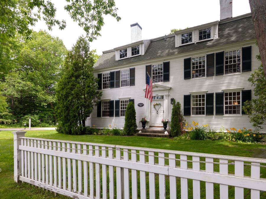 A two-story white house with black shutters, a white picket fence, and an American flag.