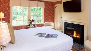 A hotel room with a large bed, a fireplace, a TV mounted above the fireplace, and a window with a view of greenery.