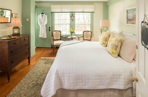 A bedroom with a large bed, a dresser, two chairs, a window with curtains, and a vase with flowers.