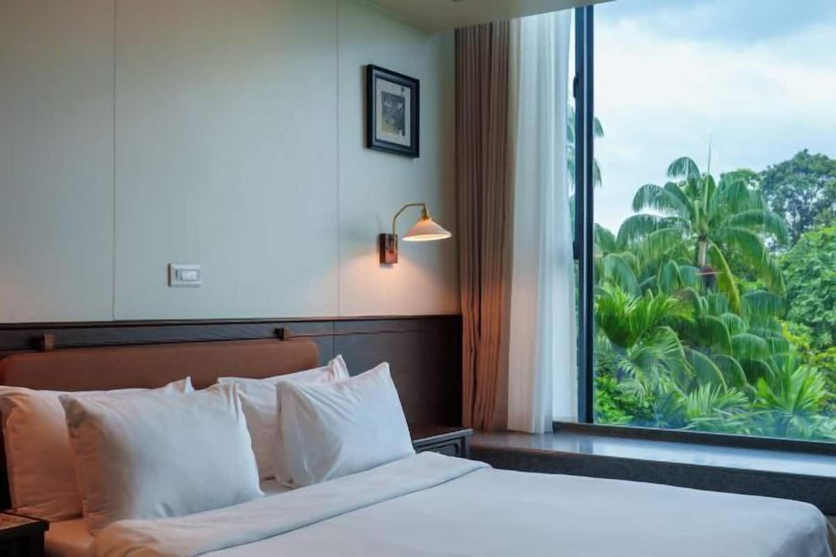 Habitación de hotel con cama, mesita de noche, lámpara de pared, un cuadro enmarcado y ventana con vistas a zonas verdes.