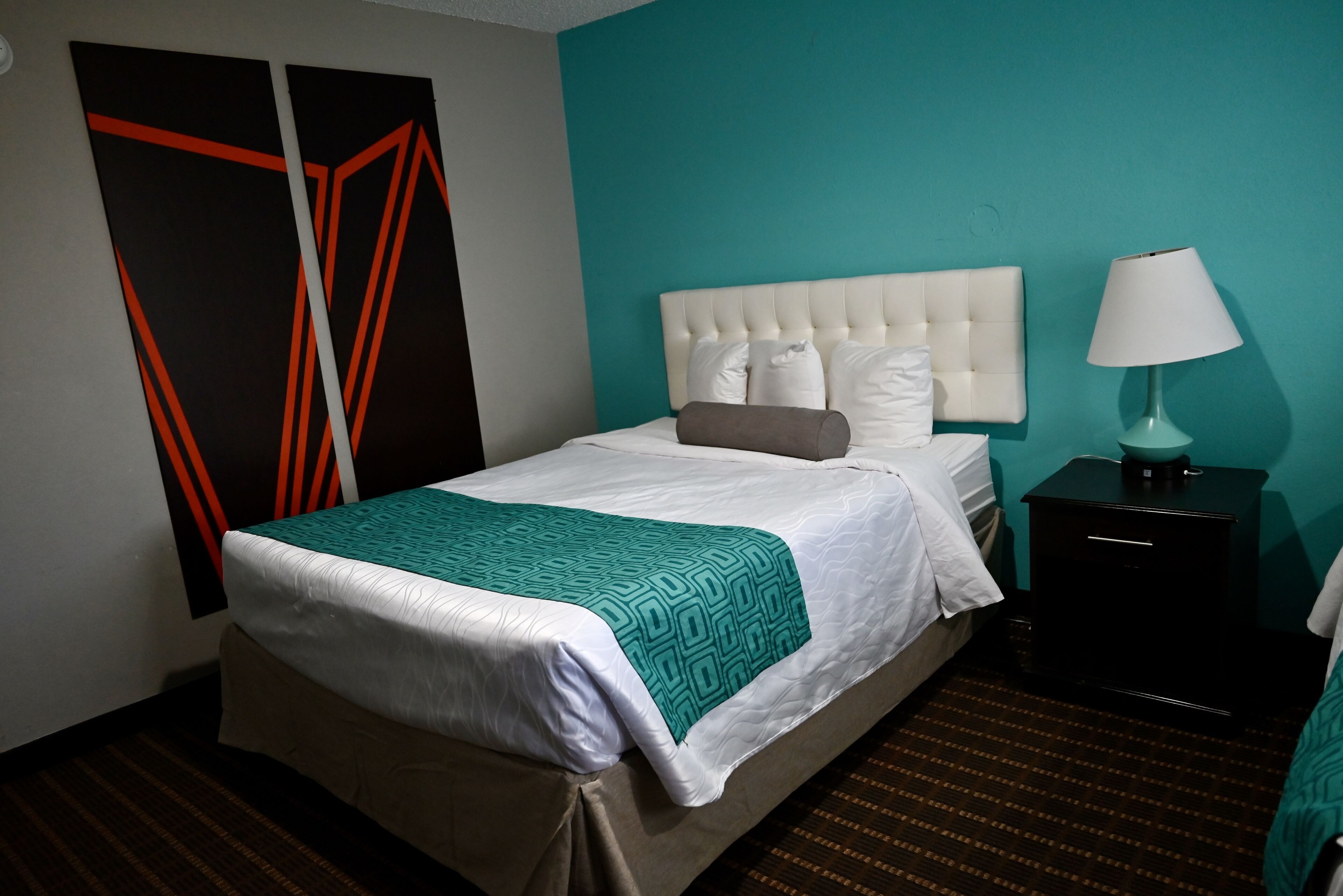 Un lit bien fait, avec une tête de lit blanche et un plaid turquoise, une lampe blanche posée sur une table de chevet noire, et un tapis à rayures.