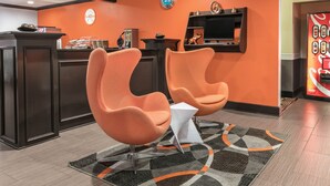 Un área de recepción con dos sillones naranjas, una mesita blanca, una alfombra estampada, un reloj y una máquina expendedora.