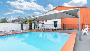Una piscina con una marquesina blanca, rodeada de sillas blancas, y un edificio de ladrillo al fondo.
